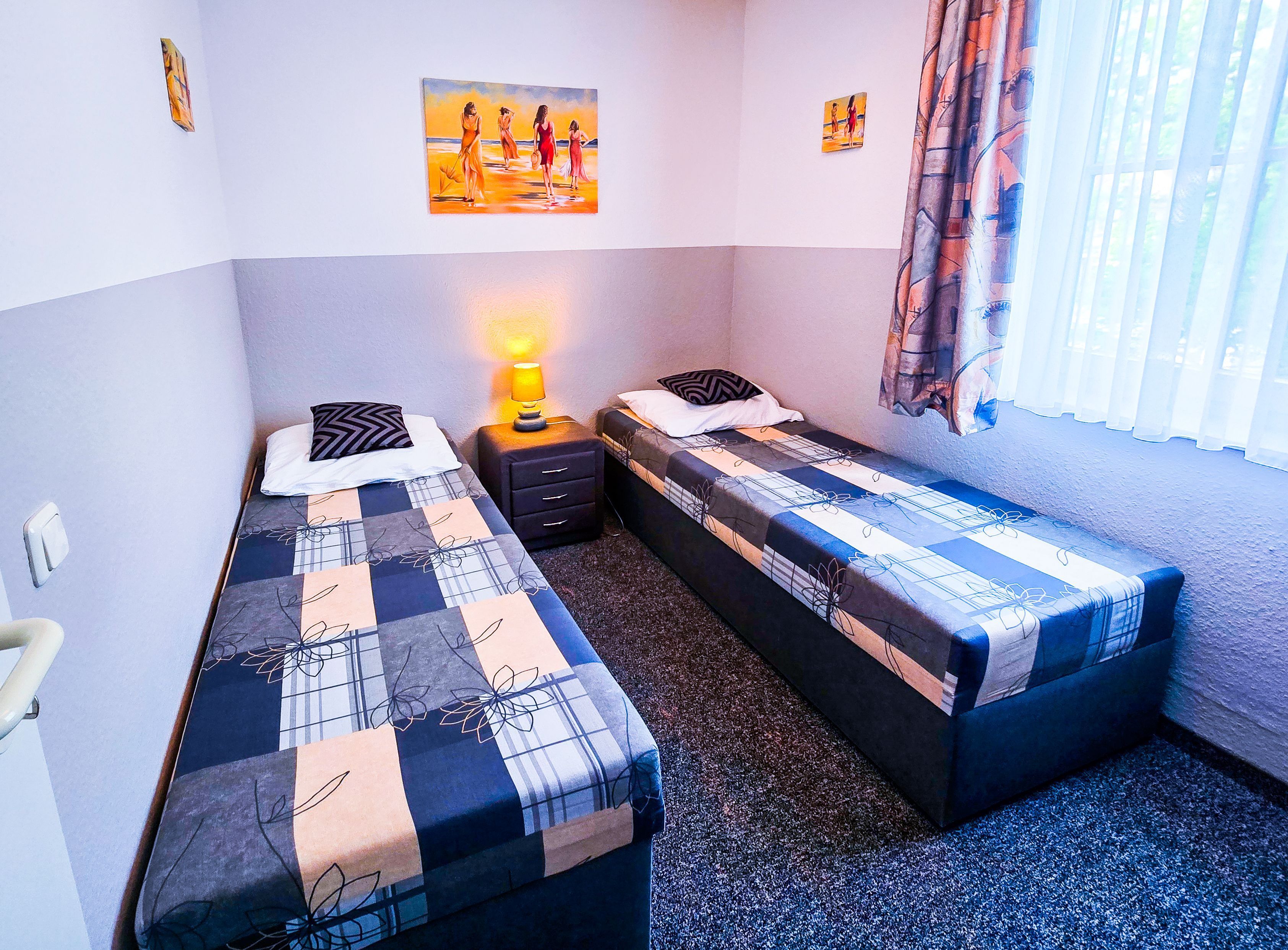 Une petite chambre propre, avec deux lits simples, une table de chevet avec une lampe et des œuvres d’art accrochées aux murs.