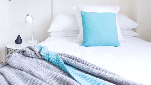 Un lit bien fait, avec une literie blanche et un seul oreiller bleu, une couverture rayée grise et bleue, une table de chevet blanche avec un vase noir et une lampe blanche.