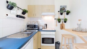 Une cuisine compacte équipée d’un évier, d’un four à micro-ondes, d’un grille-pain et d’un four. Des plantes en pot sont disposées sur le mur, et une table en bois est accompagnée de tabourets.