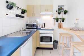 A compact kitchen with a microwave, toaster, kettle, and oven. There are potted plants, wooden utensils, and a small wooden table.