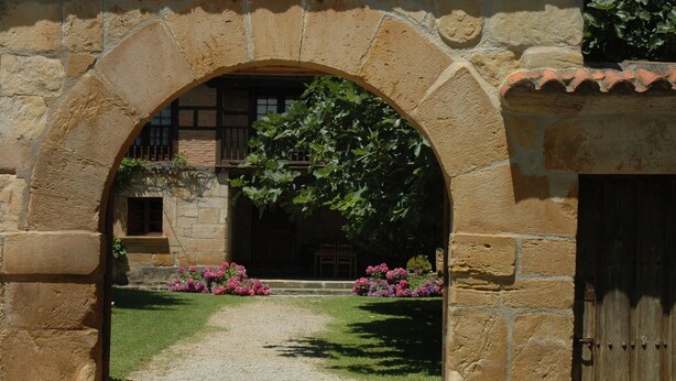 Un arc en pierre mène à un bâtiment doté d’une porte en bois et d’un petit jardin fleuri.