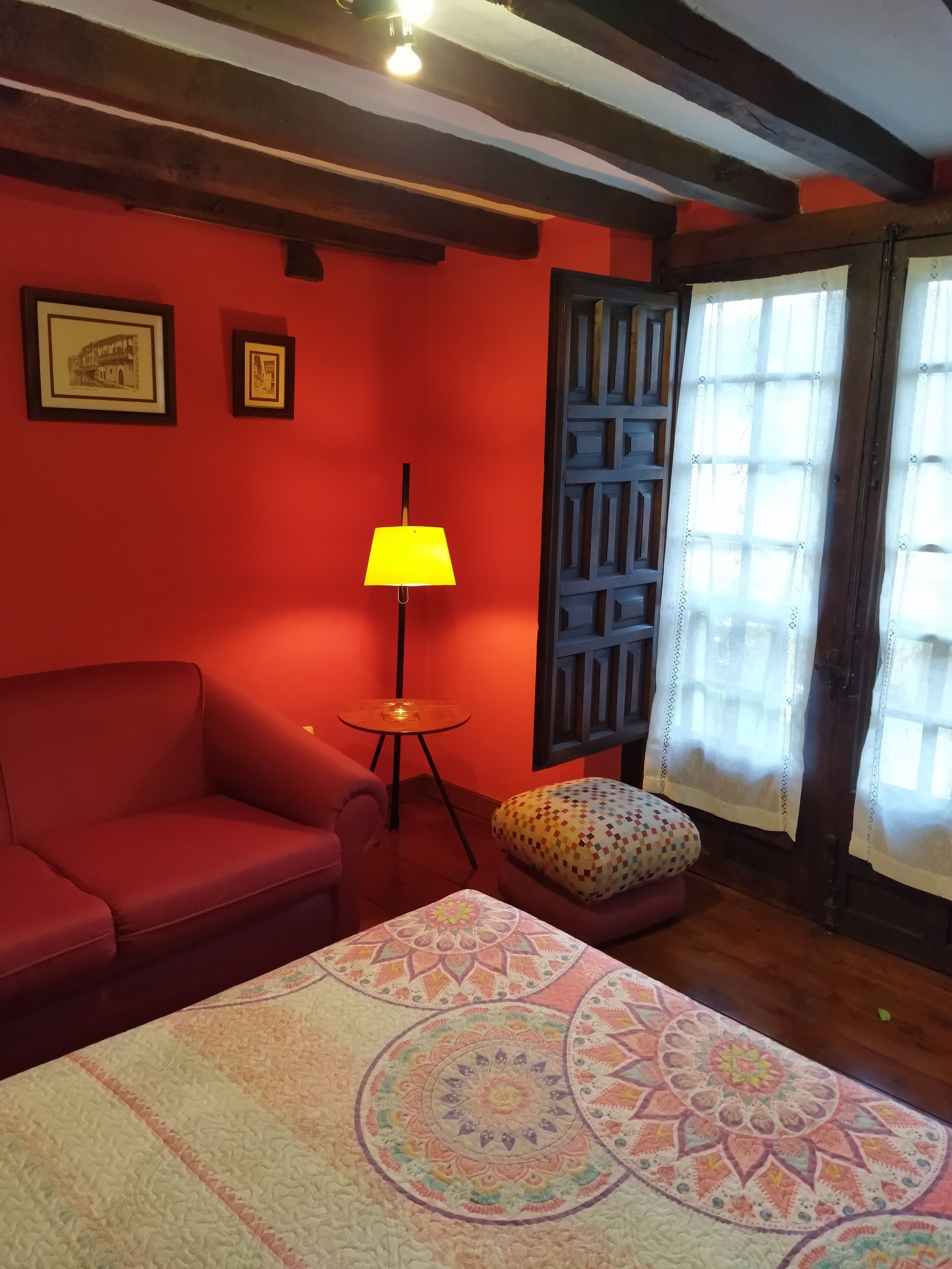 A room with a red wall, a wooden ceiling, a red sofa, a small round table with a lamp, a colorful cushion, and a patterned rug.