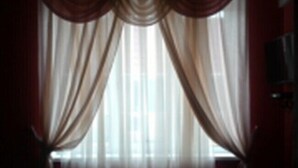 A room with a large window covered by sheer curtains, a wooden floor, and a television on the right side. - Galant Hotel (Boryspil)