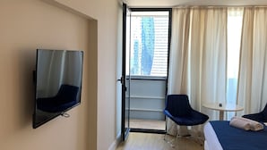 A hotel room with a bed, a TV mounted on the wall, a balcony with a view of tall buildings, and a small table with a chair. - Orbi city Davide (Batumi)