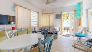 Amplia sala de estar con mesa y sillas de comedor, sofá y televisor. Cuenta con ventilador de techo, ventana con cortinas y puerta que da al exterior.