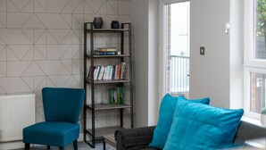 Un salon moderne avec un fauteuil turquoise, un canapé foncé agrémenté de coussins bleus, une bibliothèque et un mur orné d’un motif géométrique.