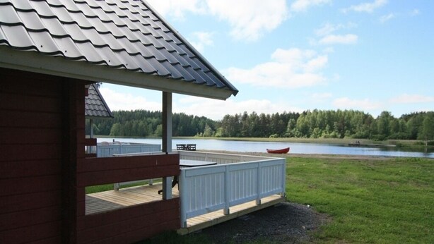 Una cabaña de madera con terraza que da a un lago, rodeada de bosque.