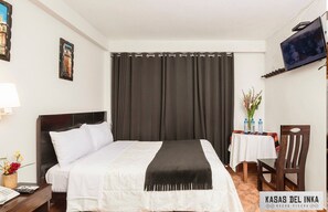 A hotel room with a bed, a desk with a lamp, a chair, a TV mounted on the wall, and a table with a vase of flowers. - "Te sentirás como en Casa, estando en Machu Picchu" (Aguas calientes)