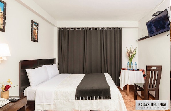 A hotel room with a bed, a desk with a lamp, a chair, a TV mounted on the wall, and a table with a vase of flowers.