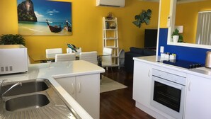 A modern kitchen with white cabinets, a stainless steel sink, and a built-in oven. There is a dining area with white chairs and a glass table. A painting of a coastal scene is on the yellow wall, and there is a small shelf with decorative items. - Golden Sands Beach House, Rose Bay,  Bowen (Bowen)