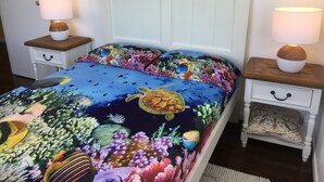 A bed with a colorful ocean-themed bedspread, two bedside tables with lamps, and a white headboard. - Golden Sands Beach House, Rose Bay,  Bowen (Bowen)