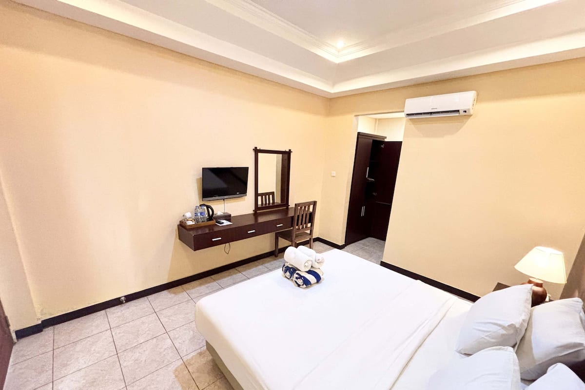 A hotel room with a bed, a desk with a flat-screen TV, a chair, and a bedside table with a lamp.