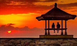 A gazebo with two people sitting under it, overlooking a rocky shoreline at sunset.