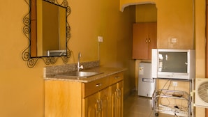 Una cocina compacta con encimera de granito, un lavamanos, un grifo de acero inoxidable, armarios de madera, un refrigerador blanco y un televisor pequeño sobre un mueble.
