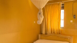Una cama individual con cabecera de madera en habitación con paredes amarillas y suelo de baldosas.