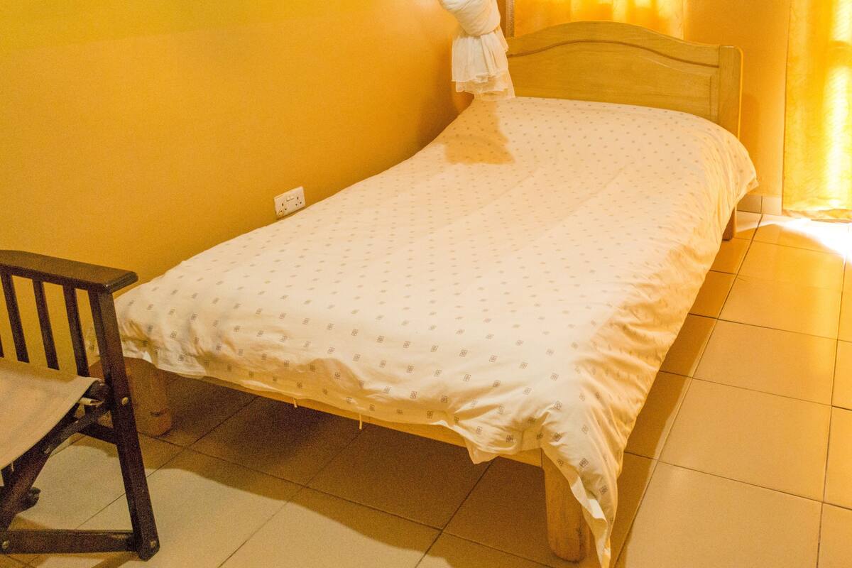 A single bed with a white bedspread and a wooden headboard in a room with yellow walls and tiled flooring.