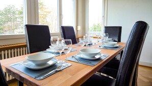 Una mesa de comedor dispuesta con platos, cuencos y copas de vino blancos, rodeada de sillas negras, con vistas a árboles a través de amplias ventanas.