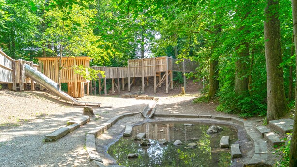 Un parque infantil con un tobogán y estructuras de madera para escalar, rodeado de árboles.
