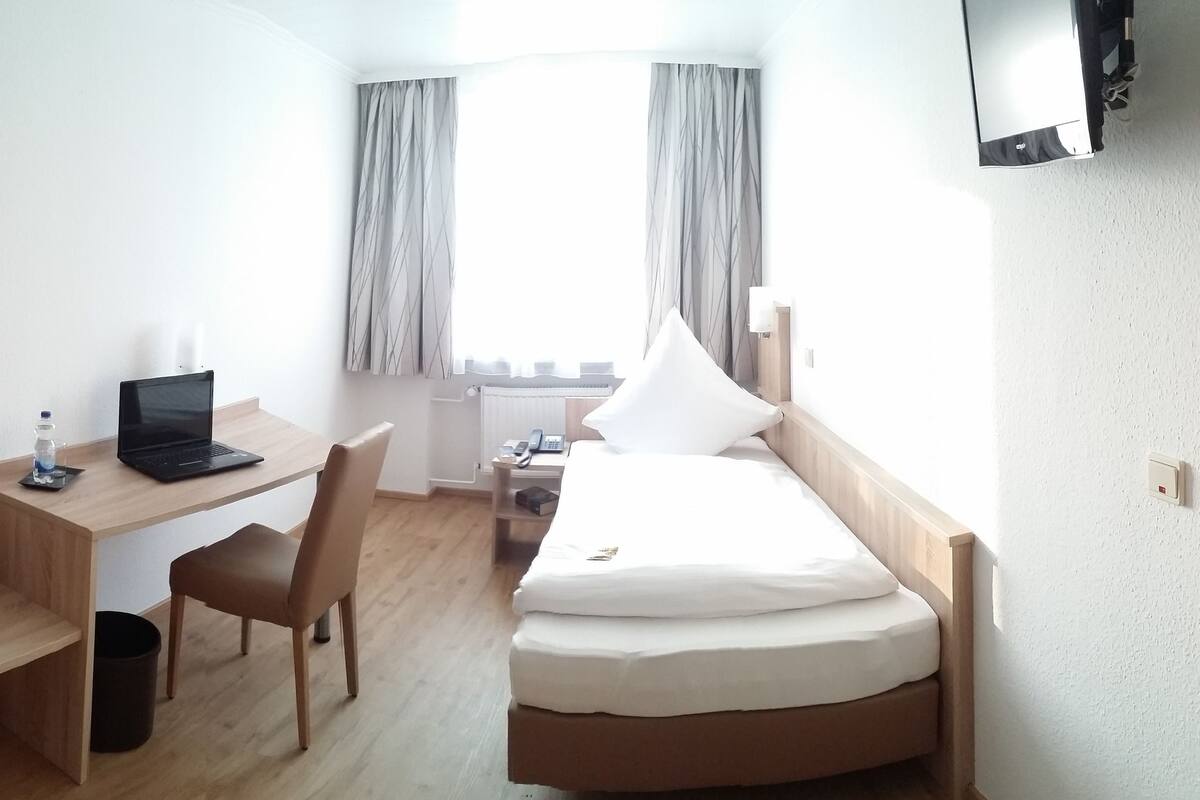 A compact hotel room with a bed, a desk with a laptop, a chair, a small table, a trash bin, and a shower area.