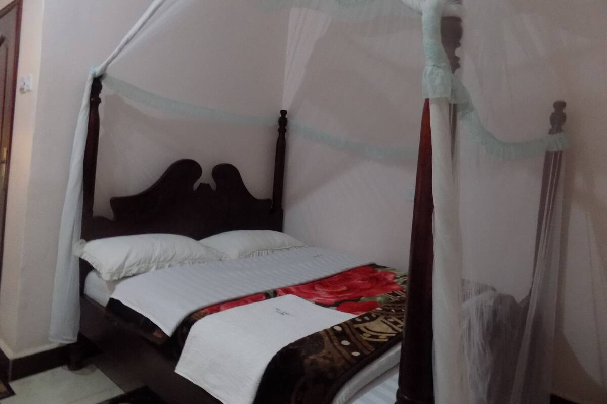 Uma cama de solteiro com cabeceira de madeira, colcha branca e manta de cor marrom e vermelha com padrão.