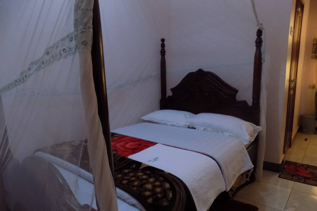 Uma cama com dossel, roupa de cama branca e um objeto decorativo vermelho sobre a cama.