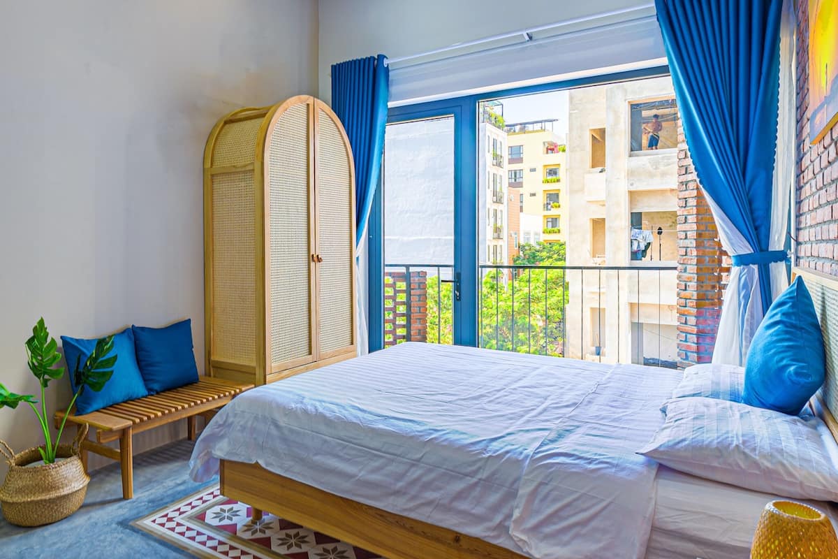 A bedroom with a bed, a bench, a rattan wardrobe, a potted plant, and a view of buildings through a window.