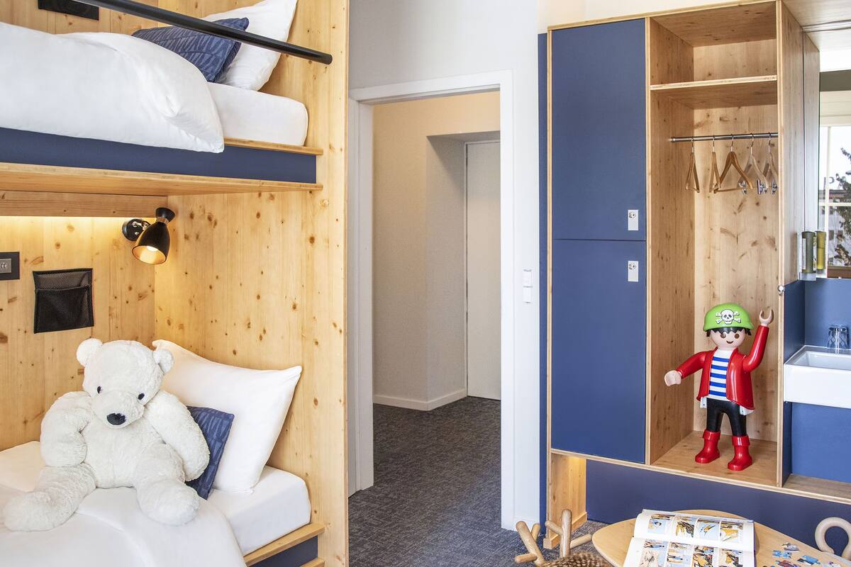 A hotel room with a bunk bed, a plush white teddy bear, a mirror reflecting a child in a pirate outfit, and a wooden wardrobe.