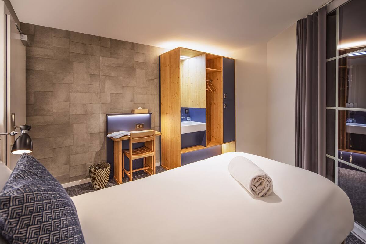 A hotel room with a bed, a desk, a chair, a small table, and a window.