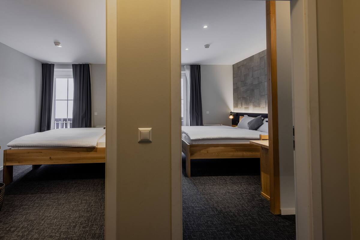 A hotel room with two beds, a large window with curtains, a wall-mounted light, and a carpeted floor.