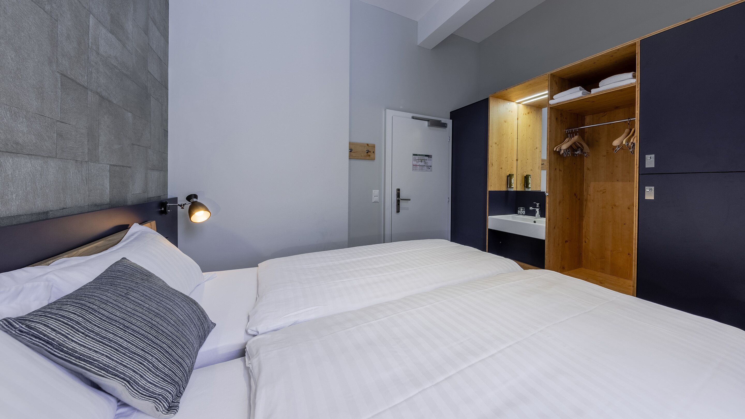 A hotel room with two beds, a wooden wardrobe, a small bathroom with a sink, and a wall-mounted light.