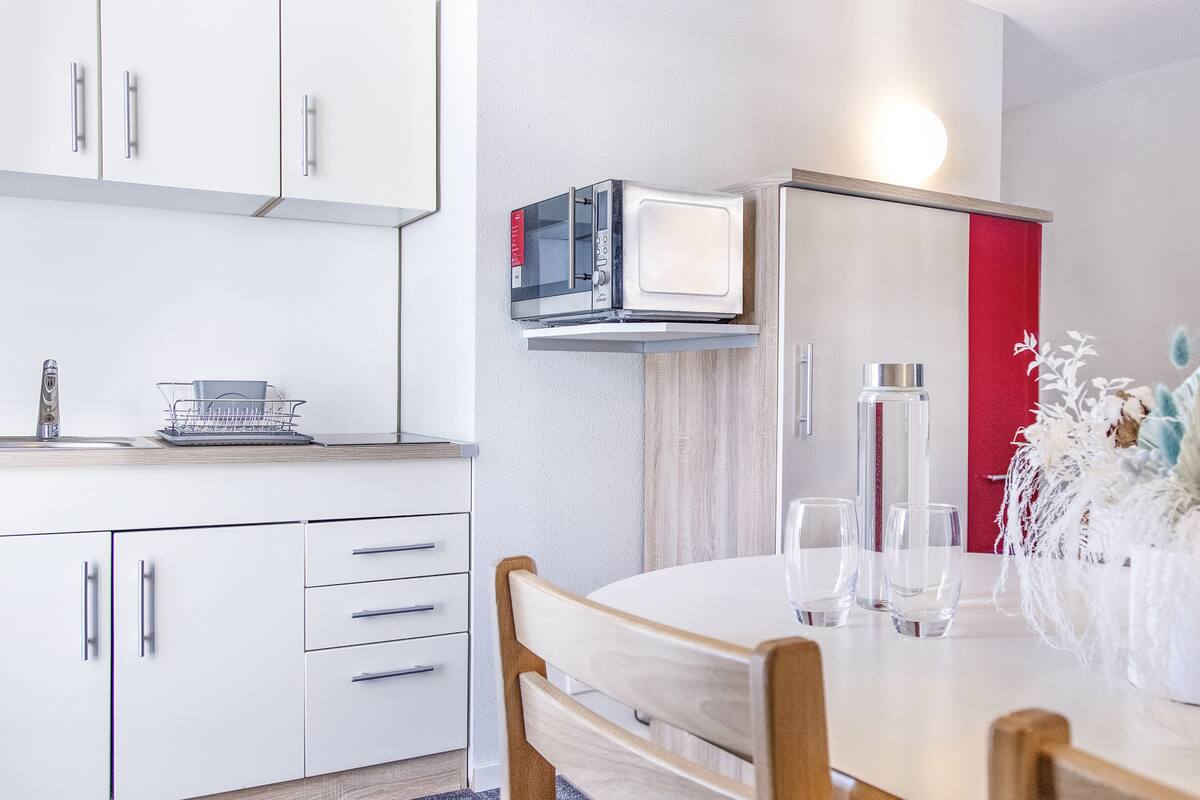 A modern kitchen with white cabinets, a microwave, and a red door. There is a dining table with chairs and a vase of dried flowers.