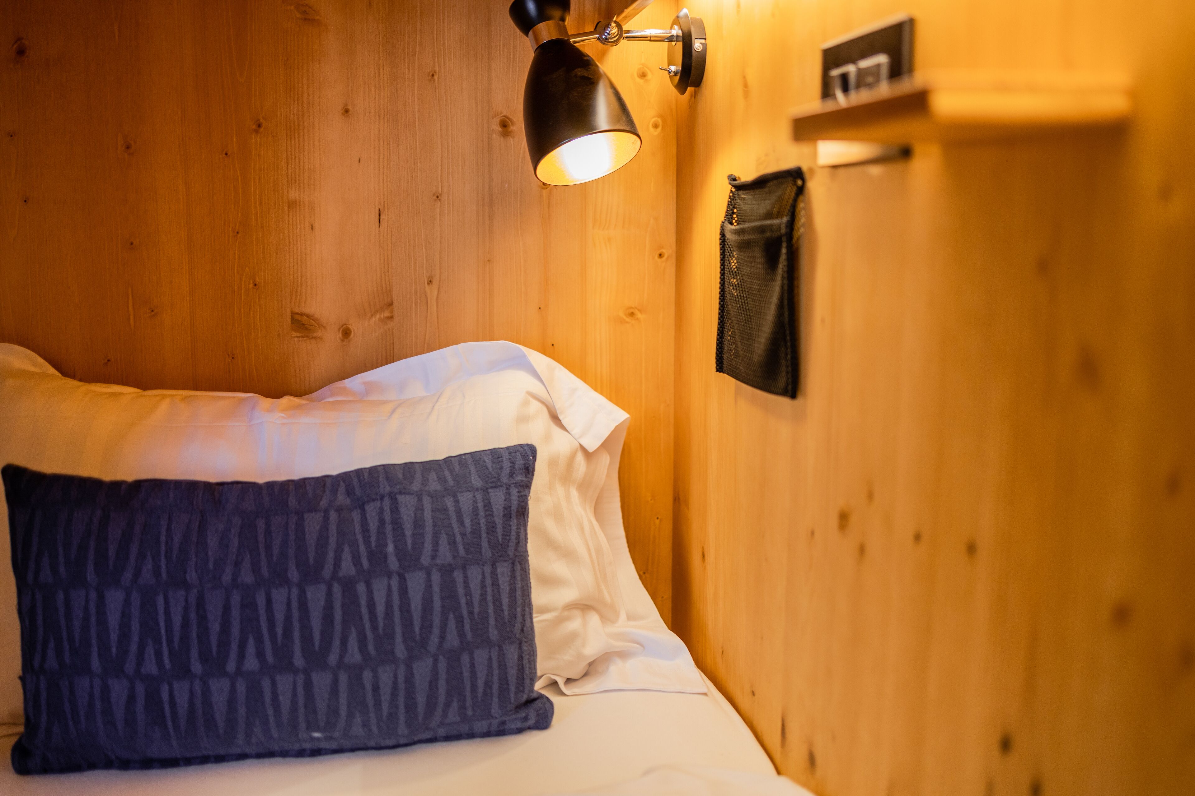 Pared de madera con una cama, un almohadón oscuro y una lámpara fijada a la pared.