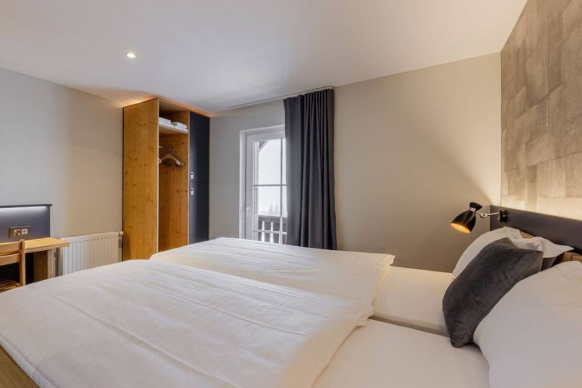 A hotel room with a bed, a wooden wardrobe, a desk with a computer, a window with curtains, and a wall-mounted lamp.