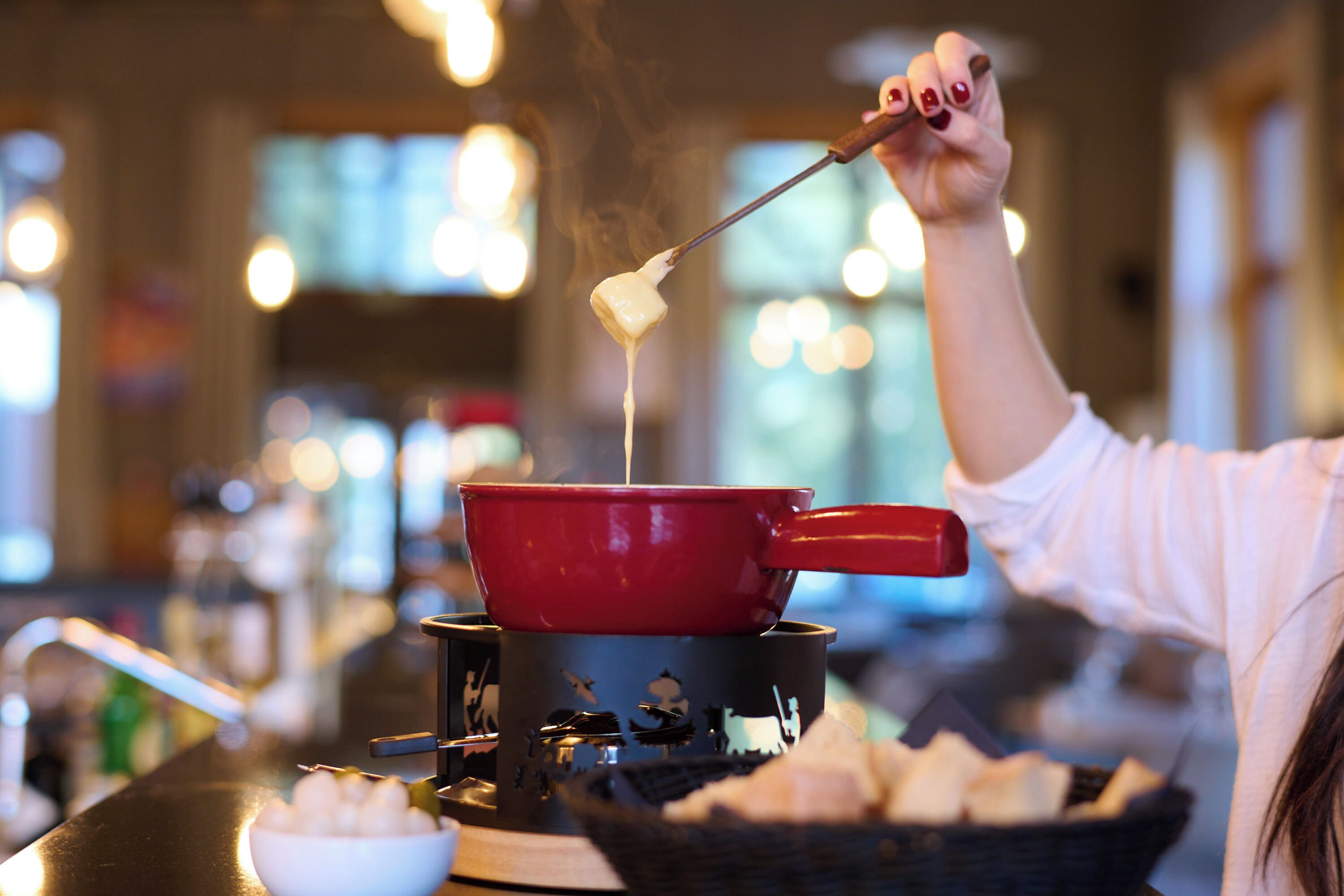 A person dipping a piece of food into a red pot of boiling liquid, likely a hot chocolate or soup, with steam rising.
