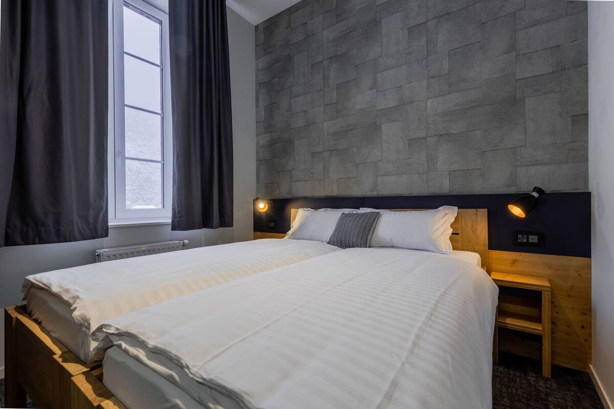 A neatly made bed with white linens, a headboard with integrated lighting, a small wooden nightstand, and a window with dark curtains.