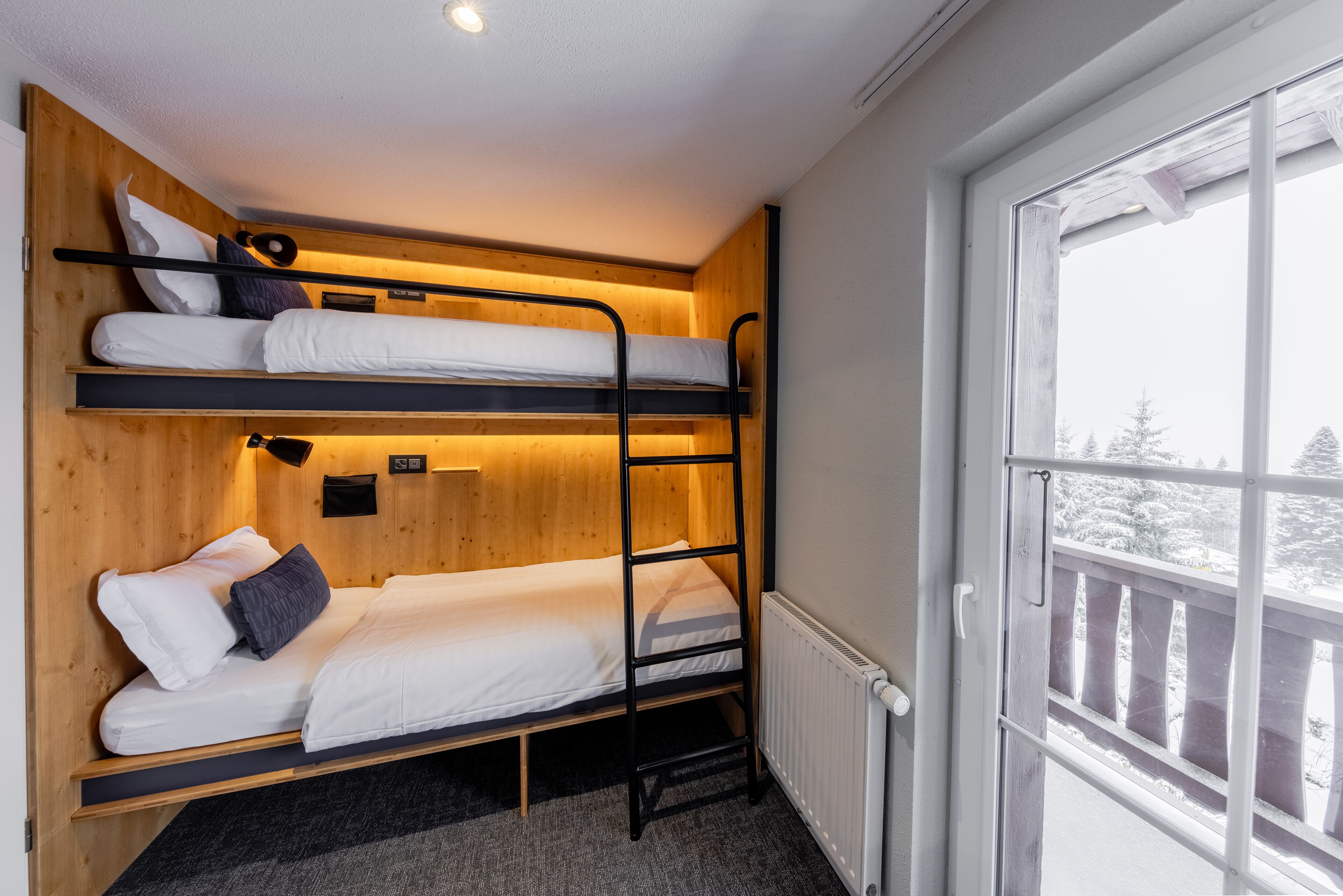 Une chambre avec un lit superposé, un mur en bois, une fenêtre et une vue sur des arbres couverts de neige.