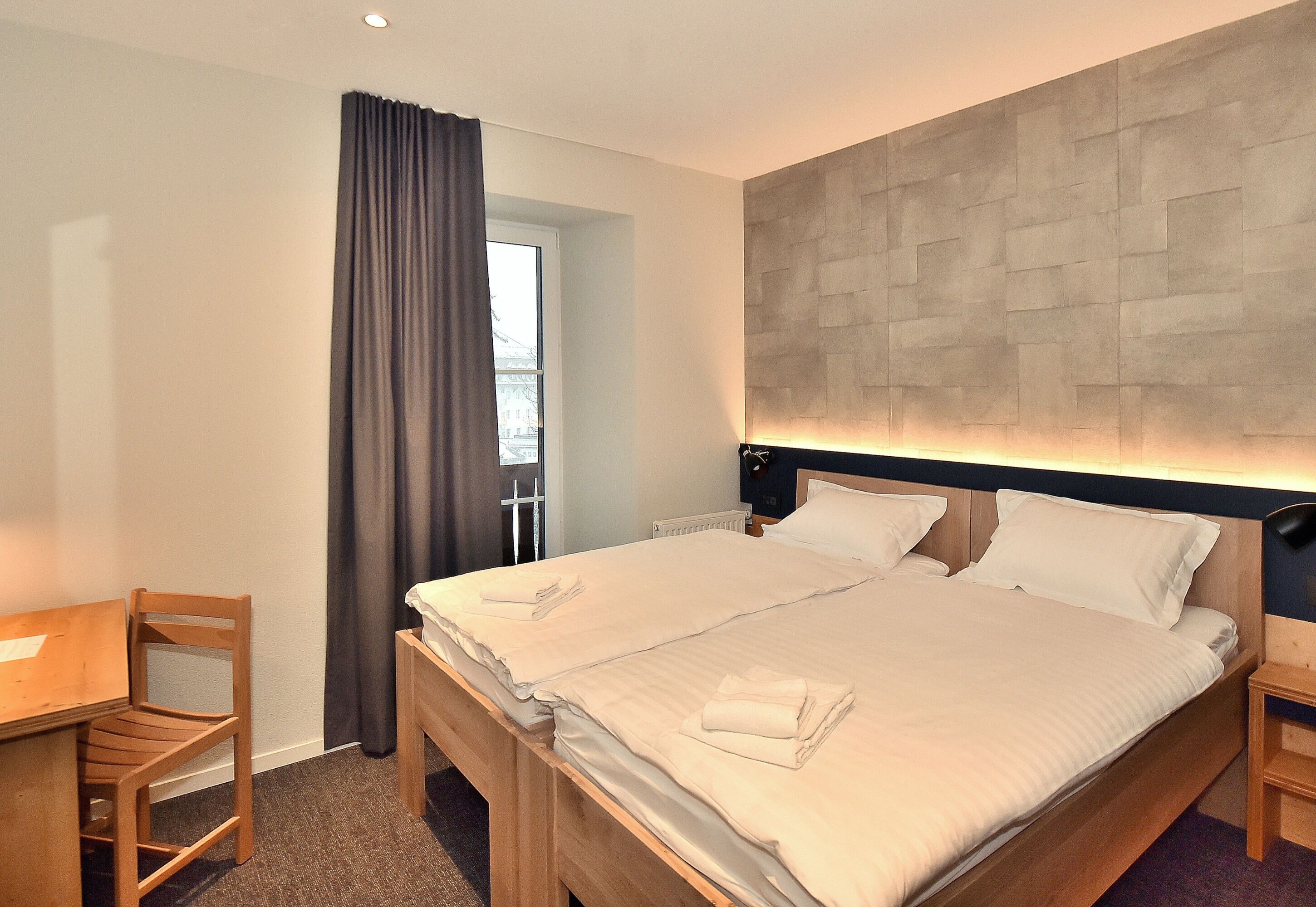 A hotel room with a double bed, a wooden desk, a chair, and a view of the city through a window with curtains.