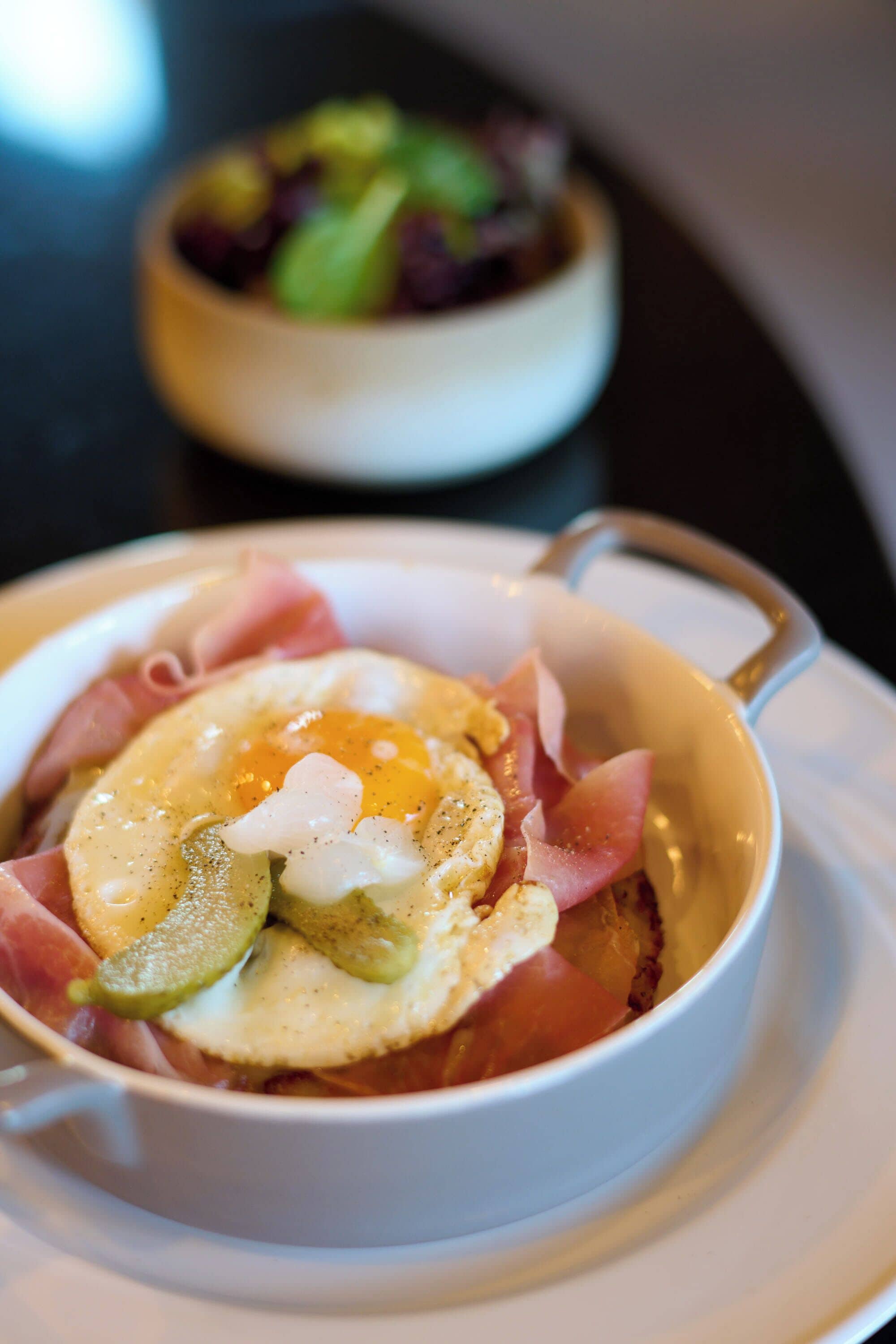 A bowl of food with a boiled egg, ham, and pickles.