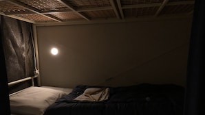 Une petite pièce faiblement éclairée, avec un lit simple, une fenêtre avec un rideau et une source de lumière visible au-dessus du lit.