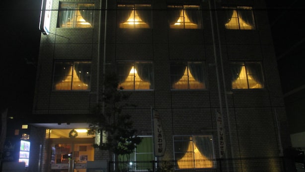 Un immeuble à plusieurs étages avec des fenêtres illuminées, une entrée bien visible et une enseigne avec des caractères japonais.