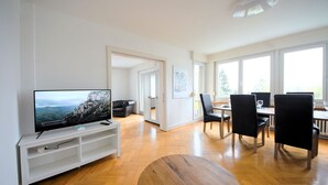 A modern living room with a flat-screen TV, wooden flooring, a dining area with black chairs, and large windows. - Sunny & Stylish 3 Bedroom Apartment, 20 min from city centre. 1st Floor. (Kreis 7)