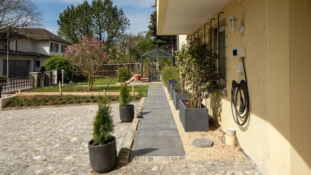 A paved pathway leading to a building with a yellow facade, a greenhouse, and potted plants along the walkway. - Sunny & Stylish 3 Bedroom Apartment, 20 min from city centre. 1st Floor. (Kreis 7)