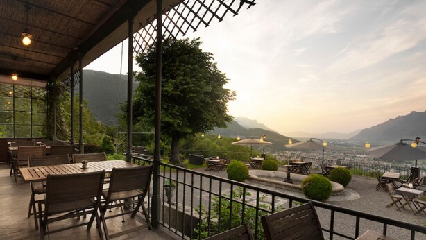 Una terraza al aire libre con mesas y sillas de madera, vista a una ciudad y montañas al fondo, y una barandilla que separa la zona de estar del paisaje.