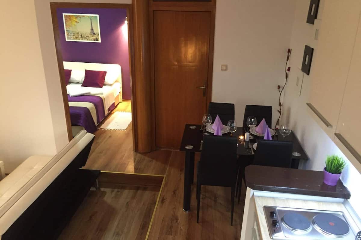 Kompakti hotellihuone, jossa on ruokailutila, violetilla tekstiilillä varustettu sänky ja pieni keittiö, jossa on liesi.
