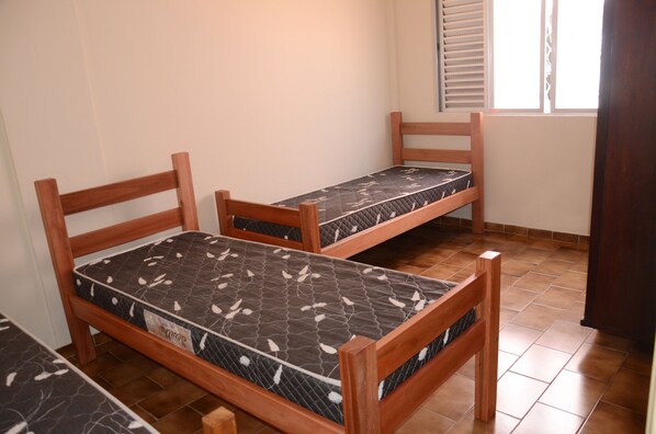 Deux lits superposés en bois, dotés de matelas à motifs, dans une pièce au sol carrelé et agréée d’une fenêtre avec des stores.