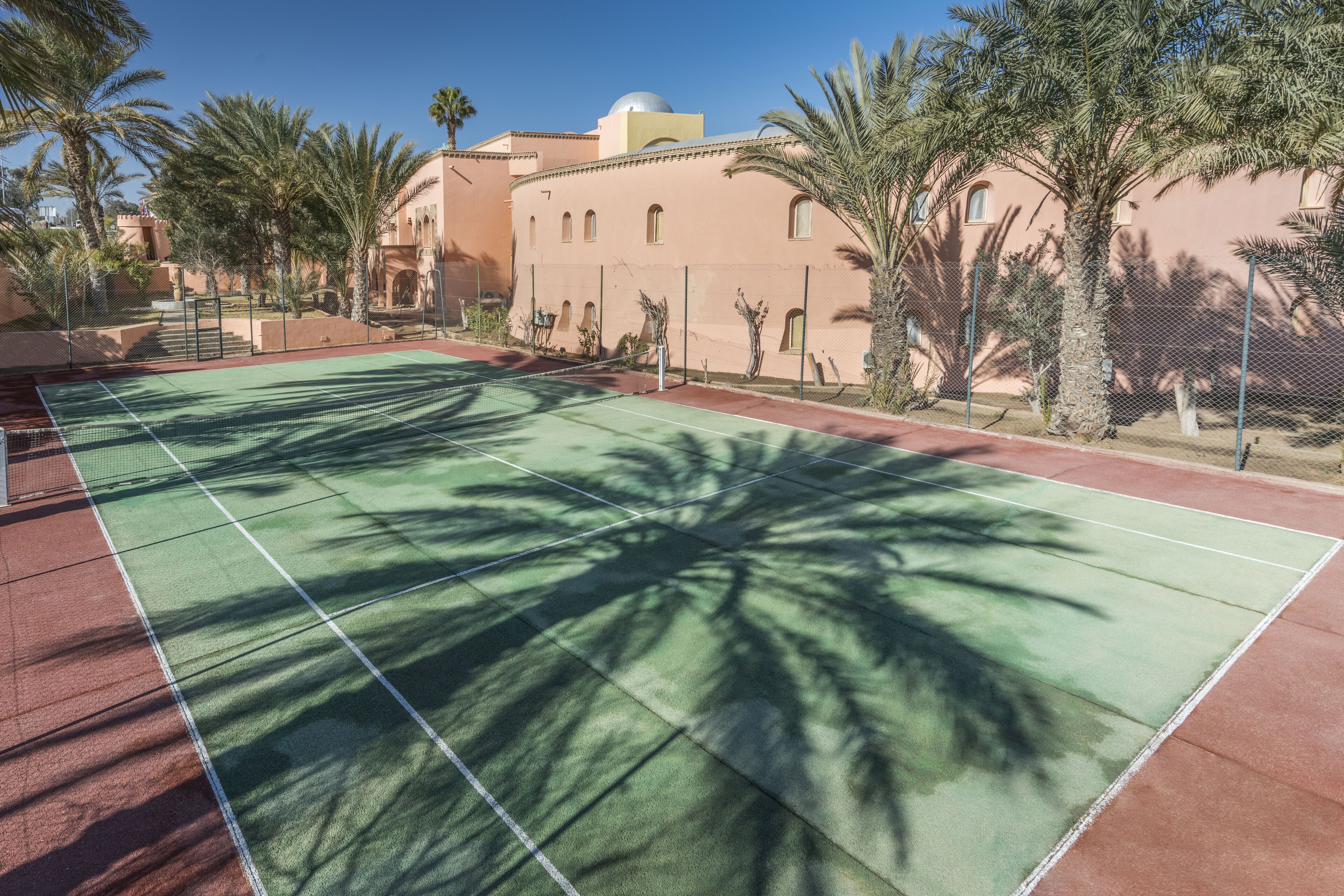 A tennis court with a net, surrounded by palm trees and a building with arched windows.