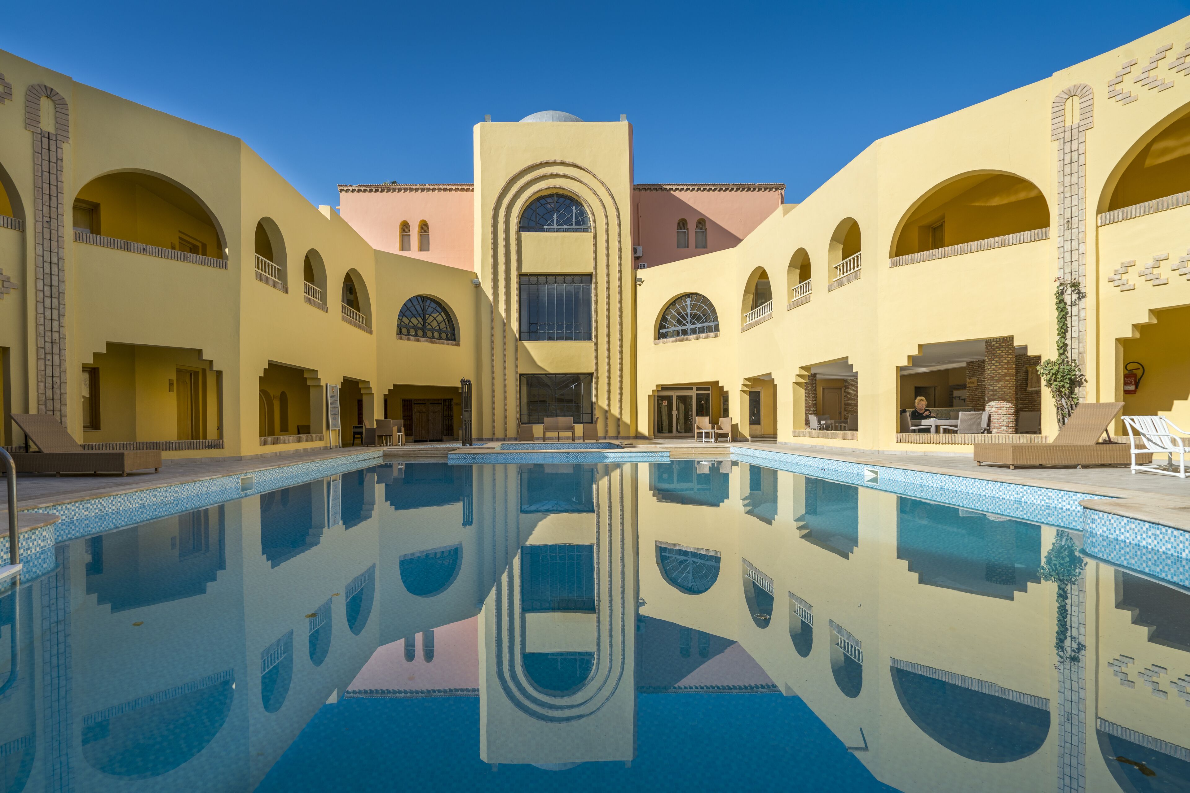 Une piscine dont l’eau est si claire qu’elle reflète parfaitement l’environnement, entourée d’un bâtiment doté de nombreuses arcades et d’une tour centrale.