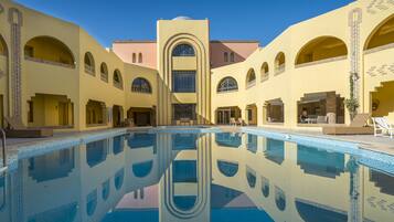 Une piscine dont l’eau est si claire qu’elle reflète parfaitement l’environnement, entourée d’un bâtiment doté de nombreuses arcades et d’une tour centrale.