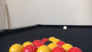 Une table de billard avec des billes et une boule blanche, un tableau encadré accroché au mur, et un panneau avec du texte.