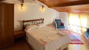 A bedroom with a wooden sloped ceiling, a bed with white bedding, a nightstand, a wardrobe, and a chair with a cushion. - Sualti Otel (Ayvalik)